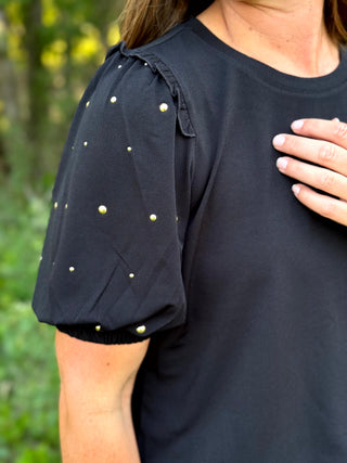 A close-up of a woman wearing a black short-sleeve top with puff sleeves and scattered gold stud embellishments, photographed outdoors with a blurred green background.