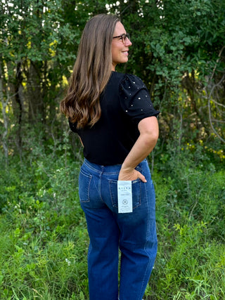 A woman wearing blue jeans with a visible Hanes paper tag on the back pocket, standing outdoors in a grassy, wooded area. She is wearing a black short-sleeve top with embellished puff sleeves and glasses.