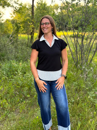 A woman wearing a black short-sleeve top with a white and black striped collar and hem, paired with blue jeans, standing outdoors in a grassy field.