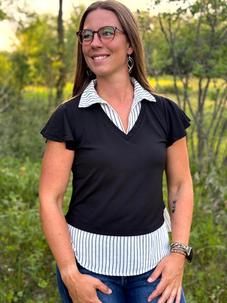 A woman wearing a black short-sleeve top with a white and black striped collar and hem detail, paired with blue jeans, standing outdoors in a green garden setting.