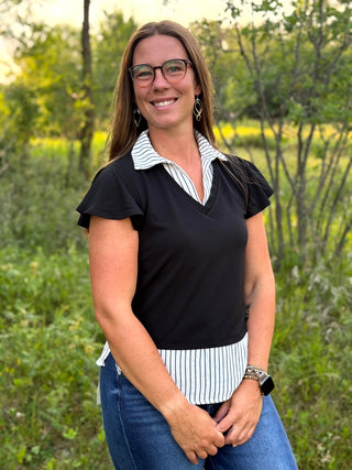 A woman wearing a black short-sleeve top with a white and black striped collar and hem detail, paired with blue jeans, standing outdoors in a grassy, wooded area.