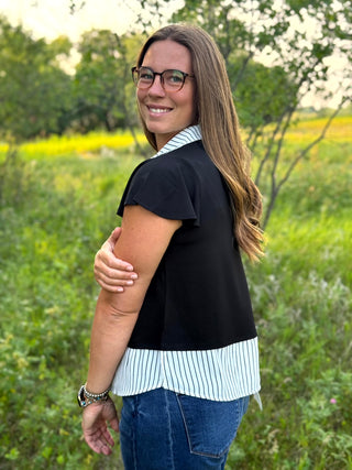 A woman wearing a black short-sleeve top with a white and black striped collar and hem, paired with blue jeans, standing outdoors in a grassy field.