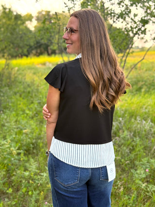 A woman wearing a black short-sleeve top with a white and black vertically striped hem, standing outdoors in a grassy field.
