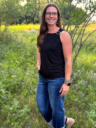 A woman wearing a sleeveless black top with a decorative silver-toned embellishment at the shoulder and blue jeans, standing outdoors in a grassy field.