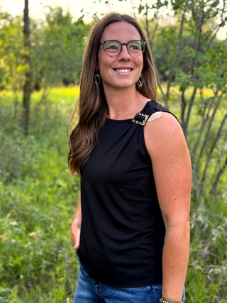 A woman with long brown hair and glasses, wearing a black sleeveless top with a decorative gold-toned embellishment on the shoulder, standing outdoors in a grassy, wooded area.
