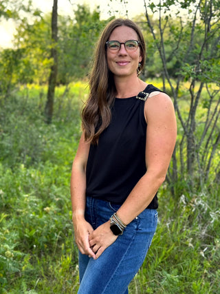 A woman with long brown hair and glasses stands outdoors in a grassy field, wearing a sleeveless black top with decorative silver embellishments on the shoulders and blue jeans.