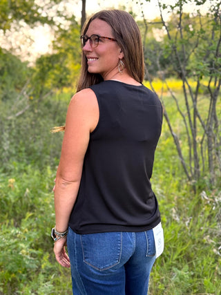 A woman wearing a sleeveless black top and blue jeans, photographed outdoors in a grassy, wooded setting.