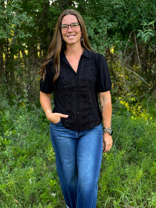A woman wearing a black short-sleeve button-up blouse with lace-up front detailing and blue jeans, standing outdoors in a grassy, wooded area.