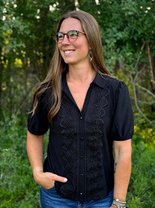 A woman with glasses and long brown hair wearing a black short-sleeve button-up blouse with black lace embroidery down the front, paired with blue jeans, standing outdoors against a green leafy background.