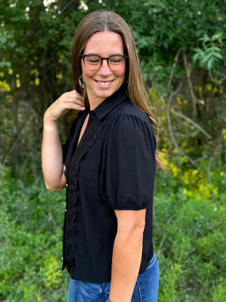 A woman wearing a black short-sleeve blouse with a scalloped lace front and puff sleeves, paired with blue jeans, standing outdoors in front of a green, blurred background.