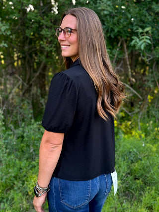 A woman with long brown hair and glasses, wearing a black puff-sleeve blouse and blue jeans, standing outdoors in front of a green leafy background.