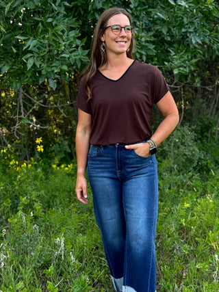 A woman wearing a dark brown v-neck t-shirt and blue jeans standing outdoors in a grassy area with green foliage.