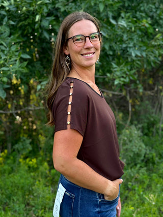A woman with long brown hair and glasses, wearing a dark brown short-sleeve top with decorative gold-toned buttons down the side and blue jeans, standing outdoors in front of a green leafy background.