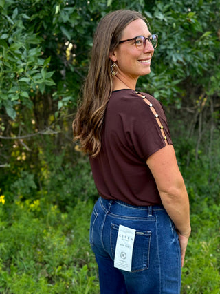 A woman wearing a dark brown short-sleeve top with decorative cutout details on the back and blue jeans, standing outdoors in front of green foliage. A white paper tag is visible on the back pocket of the jeans.