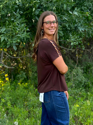 A woman wearing a dark brown short-sleeve top with decorative cutout details on the shoulders and blue jeans, standing outdoors in front of a green leafy background.