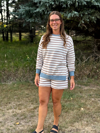 A woman wearing a matching two-piece lounge set with beige and blue horizontal stripes, standing outdoors in a grassy area with trees in the background.