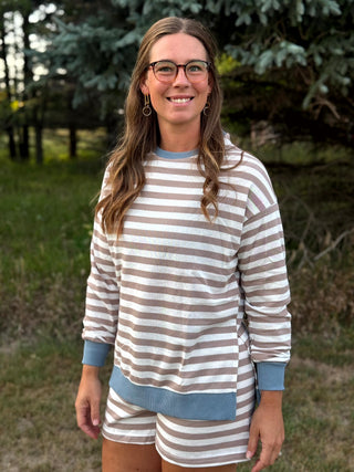 A woman wearing a matching long-sleeve striped lounge set with light blue trim, standing outdoors in a grassy area with trees in the background.