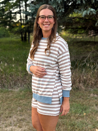 A woman wearing a brown and white striped long-sleeve tunic top with light blue trim, paired with matching striped shorts, standing outdoors in a grassy area with trees in the background.