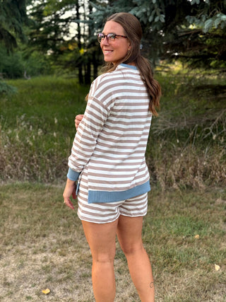 A woman wearing a matching beige and white striped long-sleeve top and shorts set with solid blue trim, standing outdoors in a grassy area with trees in the background.