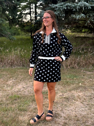 A woman wearing a black and white polka dot polo shirt and matching mini skirt stands outdoors on a grassy field with trees in the background.