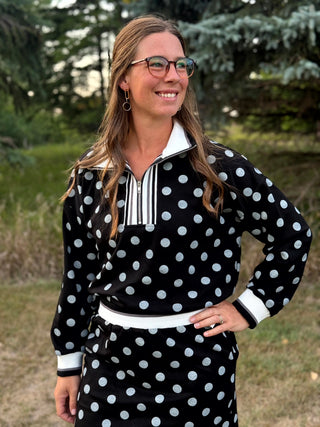 A woman wearing a black long-sleeve polo-style top with white polka dots, a white collar, and a white zipper, standing outdoors in a grassy area with trees in the background.