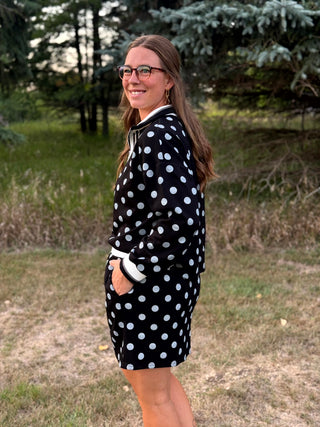 A woman wearing a black sweatshirt with white polka dots and white cuffs, standing outdoors in a grassy area with trees in the background.
