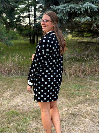 A woman wearing a black long-sleeve cardigan with white polka dots stands outdoors in a grassy field with trees in the background.