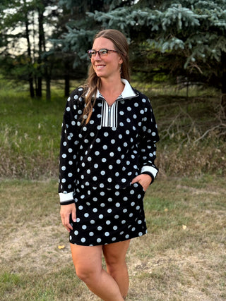 A woman wearing a black long-sleeve polo shirt dress with white polka dots and a striped collar, standing outdoors in a grassy area with trees in the background.