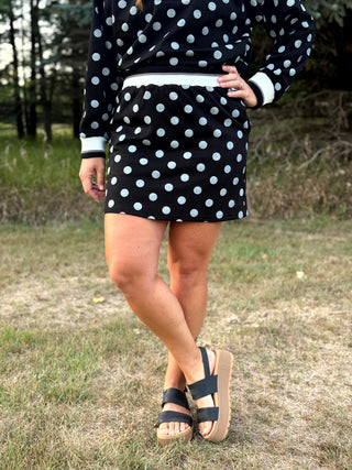 A person wearing a black long-sleeve mini dress with white polka dots and white ribbed trim, standing outdoors on grass with trees in the background.