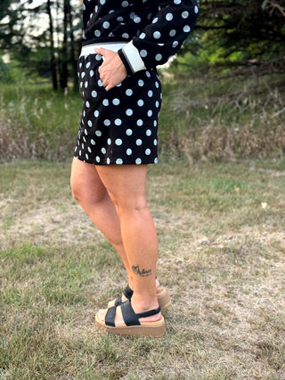 A person standing outdoors on grass wearing a black and white polka dot mini skirt with a white waistband, black platform sandals, and a matching black long-sleeve top.