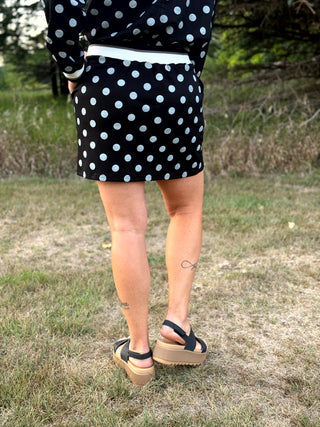 A person standing outdoors on grass wearing a black dress with white polka dots, a white belt, and black platform sandals.