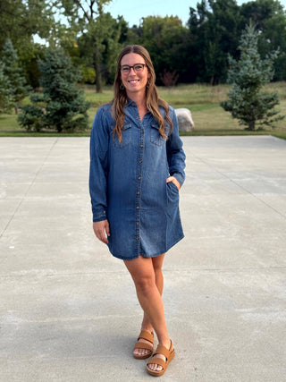 A woman wearing a blue denim button-down shirt dress and brown slide sandals, standing outdoors on a concrete patio with trees in the background.