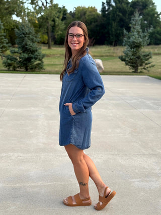 A woman wearing a blue denim long-sleeve mini dress and brown slide sandals, standing on a concrete patio with trees in the background.