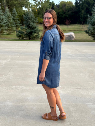 A woman wearing a blue denim shirt dress and brown slide sandals, standing outdoors on a concrete surface with trees in the background.