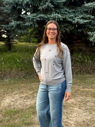 A woman wearing a light gray crewneck sweatshirt with small brown embroidered details and blue jeans, standing outdoors in front of evergreen trees and grass.