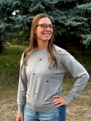 A woman wearing a light gray crewneck sweatshirt with small embroidered football motifs and blue jeans, standing outdoors in front of trees.