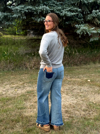 A woman wearing a light gray long-sleeve top and blue flare jeans with a dark navy pocket, standing outdoors on a grassy lawn with trees in the background.