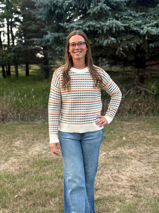 A woman wearing a cream knit sweater with horizontal multicolor stripes and blue jeans, standing outdoors in a grassy area with trees in the background.