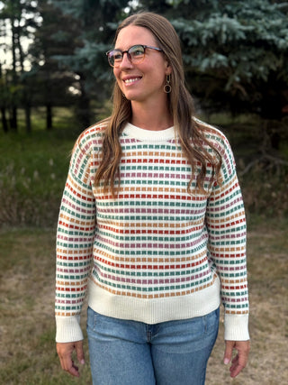 A woman wearing a cream knit sweater with a colorful horizontal stripe pattern of small squares, paired with blue jeans, standing outdoors in front of trees.
