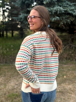 A woman wearing a cream knit sweater with a colorful geometric grid pattern in red, green, orange, and blue, standing outdoors in a park.