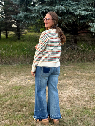 A woman wearing a cream knit sweater with a multicolor geometric pattern and blue flare jeans, standing outdoors on a grassy lawn with trees in the background.