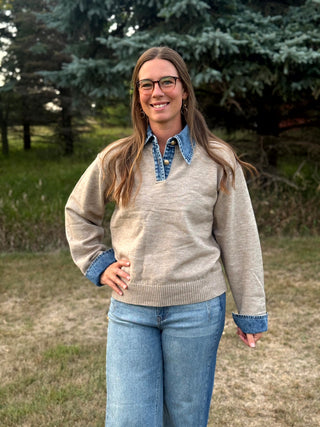 A woman wearing a beige knit sweater with blue denim trim on the collar, cuffs, and hem, paired with blue jeans, standing outdoors in a grassy area with trees in the background.