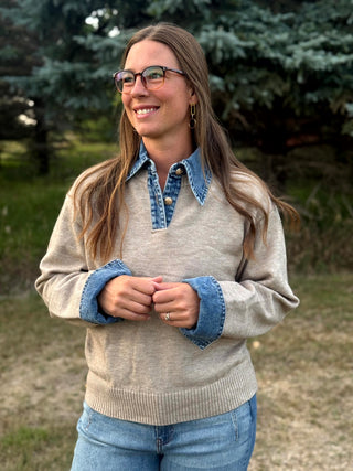 A woman wearing a beige knit sweater with blue denim collar and cuff accents, layered over a blue denim button-up shirt, standing outdoors in front of trees.