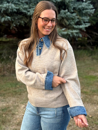 A woman wearing a beige knit sweater with a blue denim collar and matching blue denim cuffs, paired with blue jeans, standing outdoors in front of green trees.
