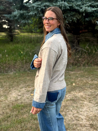 A woman wearing a light beige knit cardigan over a blue denim shirt and blue jeans, standing outdoors in a grassy area with trees in the background.