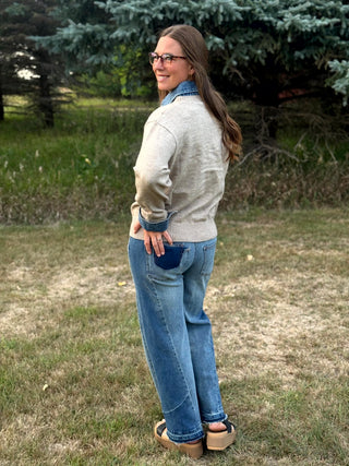 A woman wearing a light beige knit sweater, blue jeans, and brown platform sandals stands outdoors on a grassy lawn with trees in the background.
