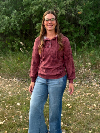 A woman wearing a maroon long-sleeve hooded sweatshirt with a subtle tonal pattern and blue jeans, standing outdoors in front of a green wooded background.
