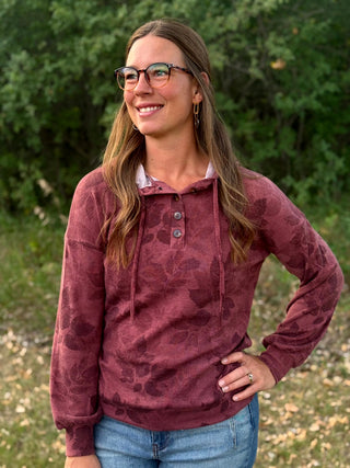 A woman wearing a dusty rose long-sleeve henley hoodie with a tonal abstract pattern, blue jeans, and glasses, standing outdoors in front of a green forest background.