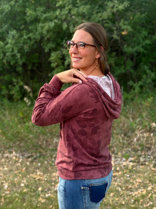 A woman wearing a maroon hooded sweatshirt with a tonal floral pattern and blue jeans, standing outdoors in front of a green wooded background.
