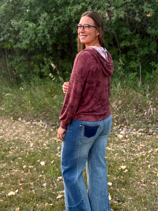 A woman wearing a maroon long-sleeve hooded sweatshirt with a tonal floral pattern and blue jeans, standing outdoors in a grassy area with trees in the background.